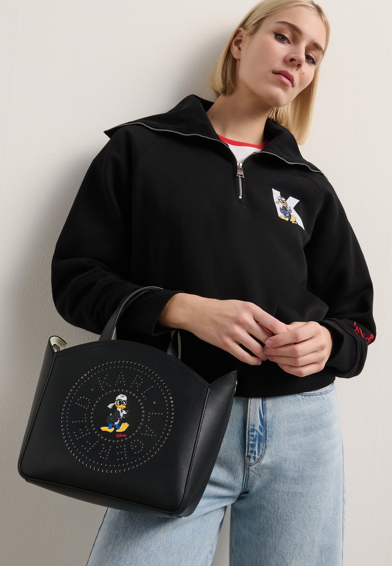 Bolsa preta feita de pele com um design circular decorado com rebites e um motivo central de personagem. A bolsa tem bordas arredondadas e alças curtas.
