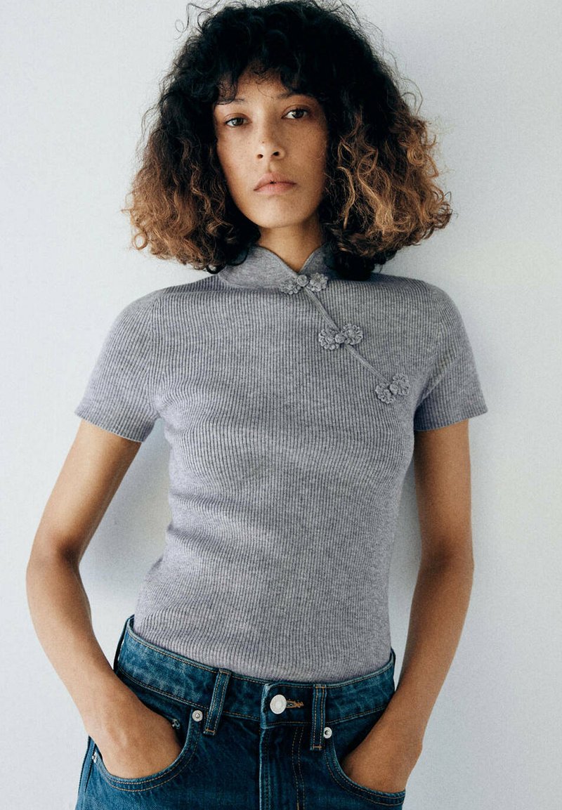 Jeune personne aux cheveux bouclés mi-longs porte un haut gris côtelé à manches courtes et un jean bleu, les mains dans les poches, sur un fond uni.