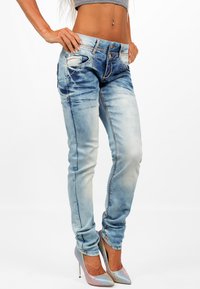 Jeans in denim azzurro chiaro con un design aderente, leggeri segni di usura, cuciture gialle e numerose tasche. Abbinati a tacchi alti iridescenti.
