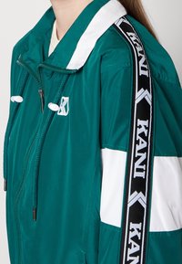 Turkusowa nylonowa kurtka z białymi akcentami. Posiada regulowane sznurki, branding KANI na rękawach oraz zamek błyskawiczny z przodu.