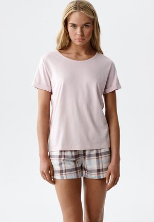 Młoda kobieta o długich blond włosach, ubrana w jasnoróżową koszulkę z krótkim rękawem i kraciaste szorty, stoi na tle jednolitego, jasnego tła.