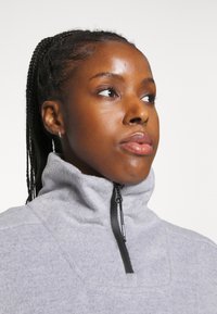 Camisa de fleece cinza claro com zíper de um quarto, destacando um colarinho alto, zíper preto e textura macia. Detalhe de costura minimalista no peito.