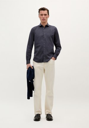 Cămașă bleu închis cu mânecă lungă, butonată, cu guler; pantaloni bej; ținând un pulover bleu închis; pantofi negri; design simplu pe un fundal deschis.