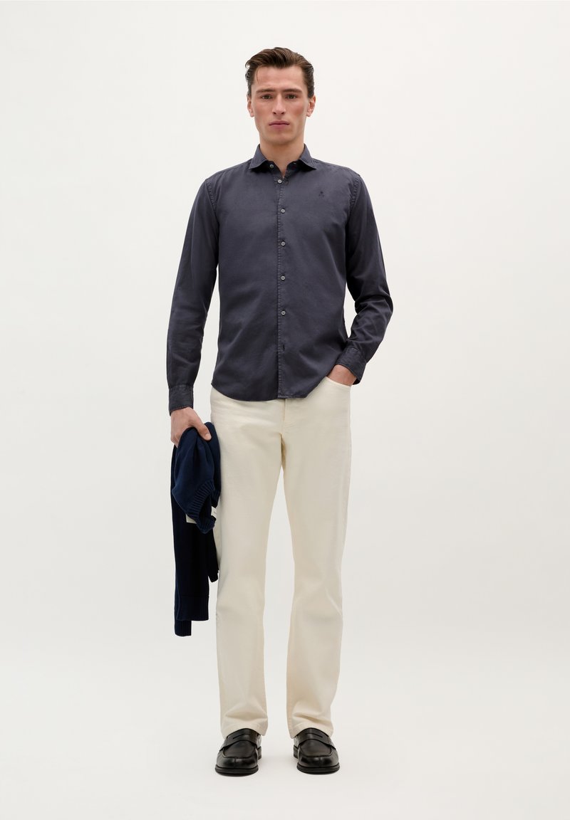 Cămașă bleu închis cu mânecă lungă, butonată, cu guler; pantaloni bej; ținând un pulover bleu închis; pantofi negri; design simplu pe un fundal deschis.