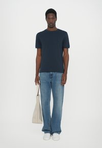Marineblå kortermet t-skjorte med rund hals, sammen med lyseblå vide jeans og hvite sneakers, holder en lys grå totebag.