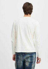 Langærmet cremet skjorte lavet af blødt stof, med rund halsudskæring, parret med blå slidte jeans og synlig syning på lommerne.