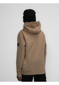 Sudadera color marrón claro con un gran patrón de logotipos sutil, capucha con cordón y puños de canalé. Un parche negro acentúa la manga izquierda.