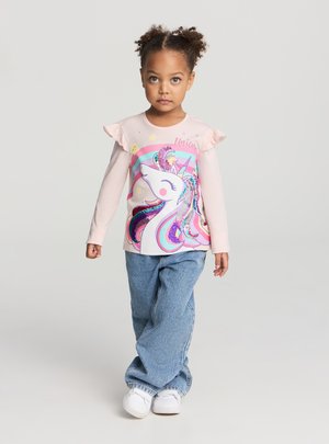 Młoda dziewczyna stojąca ze skrzyżowanymi nogami, ubrana w różową bluzę z jednorożcem, niebieskie dżinsy i białe trampki na jasnym tle.