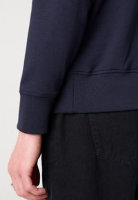 Marineblauwe sweatshirt met ribbelmanchetten en -zoom, gemaakt van een zachte, gestructureerde stof. De focus ligt op de mouw en hand tegen donkere jeans.