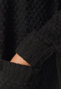 Ruka uvnitř žebrované kapsy černého pleteného svetru s texturovanými vzory.