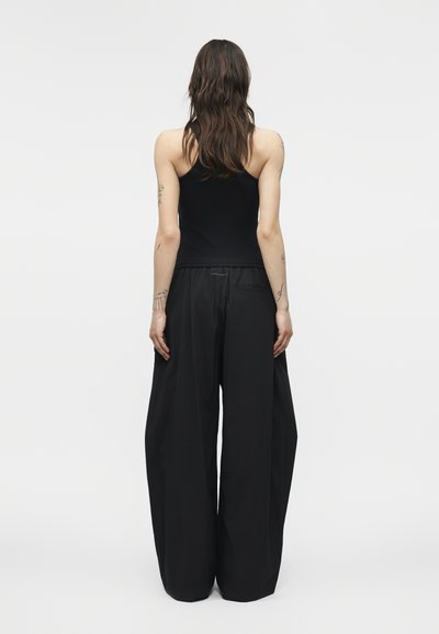 Γυναίκα με μακριά μαλλιά, φορώντας μαύρο τοπ χωρίς μανίκια και μαύρο παντελόνι με φαρδιά μπατζάκια, στέκεται με την πλάτη προς τον θεατή.