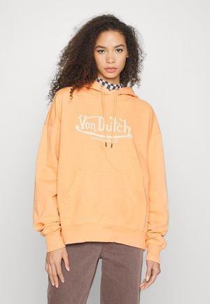 Oransje hettegenser med frontlomme og snøring i hetten, med "Von Dutch Originals"-logo i hvitt. Brukt med brune bukser.