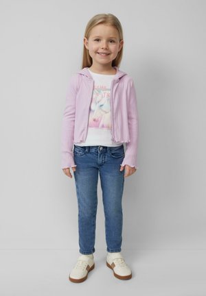 Jasnoróżowy żebrowany jacket na zamek, biały t-shirt z grafiką jednorożca, niebieskie dopasowane dżinsy oraz białe sneakersy z brązowymi podeszwami.