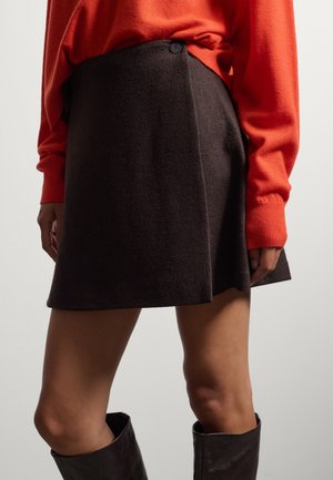 Frau trägt einen dunkelbraunen Woll-Mini-Wickelrock mit einem schwarzen Knopf, einen orangefarbenen Pullover mit langen Ärmeln und schwarze kniehohe Stiefel.