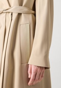 Manteau beige à manches longues avec une ceinture nouée à la taille et un gros plan d'une main pendant naturellement sur le côté.
