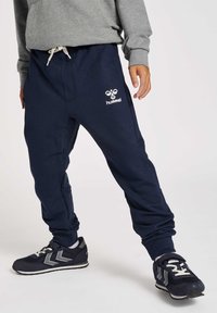 Navy-Trainingshosen mit elastischem Bund, seitlichen Taschen und weißem "hummel"-Logo. Getragen mit navy- und grauen Sportschuhen mit kontrastierenden Streifen.