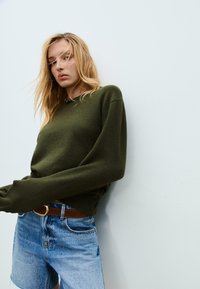 Młoda kobieta z blond włosami, w oliwkowym swetrze i dżinsowych szortach, opiera się o jasnoszary mur.