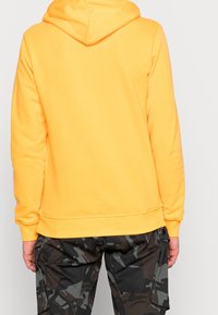 Żółta bluza z kapturem i prążkowanymi mankietami oraz dolnym wykończeniem, z regulowanym kapturem. Połączona z ciemnymi spodniami cargo w kamuflażu.