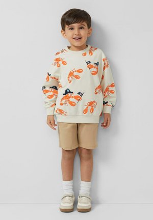 Uśmiechnięty chłopiec w kremowej bluzie z nadrukiem pomarańczowego homara i pirackiego kapelusza, beżowych szortach, białych skarpetkach i beżowych trampkach.