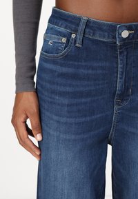 Σκούρο μπλε τζιν παντελόνι με ψηλή μέση, κλασικό σχέδιο πέντε τσεπών και ελαφριά ραφή. Το χέρι resting στην περιοχή της μέσης.