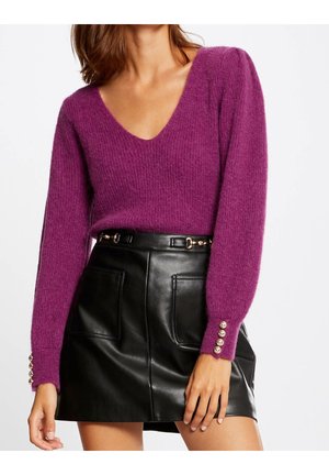 Kvinde iført en lilla V-udskæring, blød sweater med guldknapper på ærmerne og en sort lædernederdel med frontlommer og bælte detaljer.