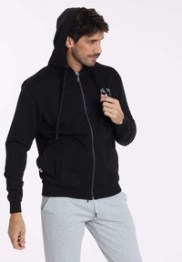 Sweat à capuche noir avec fermeture éclair et cordon de serrage. Présente un patch logo subtil et des poignets côtelés, associé à un pantalon de survêtement gris clair.