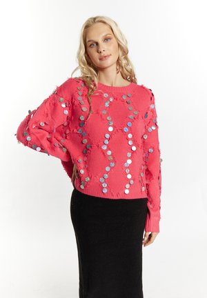 Roze gebreide trui met pailletten in een ruitpatroon en rafelige randen, gecombineerd met een zwarte getailleerde rok. Zachte textuur, losse pasvorm.