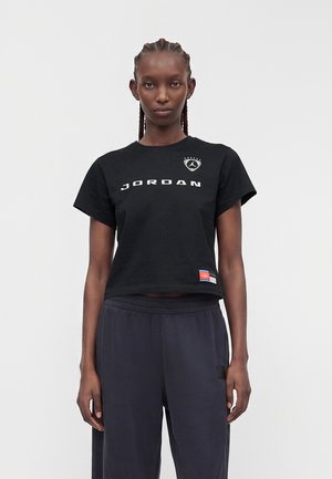 Personne grande avec des cheveux tressés portant un t-shirt Jordan noir et un pantalon ample foncé, debout sur un fond clair uni.