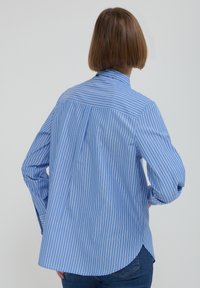 Person with short brown hair wears a blue and white vertical striped shirt and blue jeans, facing away against a plain background.