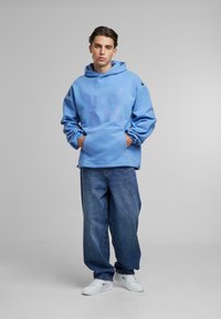 Hoodie azzurro con tasca frontale, caratterizzato da un design grafico sbiadito, abbinato a jeans blu scuri a vestibilità larga e sneakers bianche.