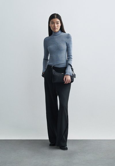 Sieviete, kas ģērbusies pieguļošā zilo svītrainajā augstajā kaklā un brīvajās melnās svītrainajās biksēs, tur mazu melnu roku somu uz vienkārša pelēka fona.