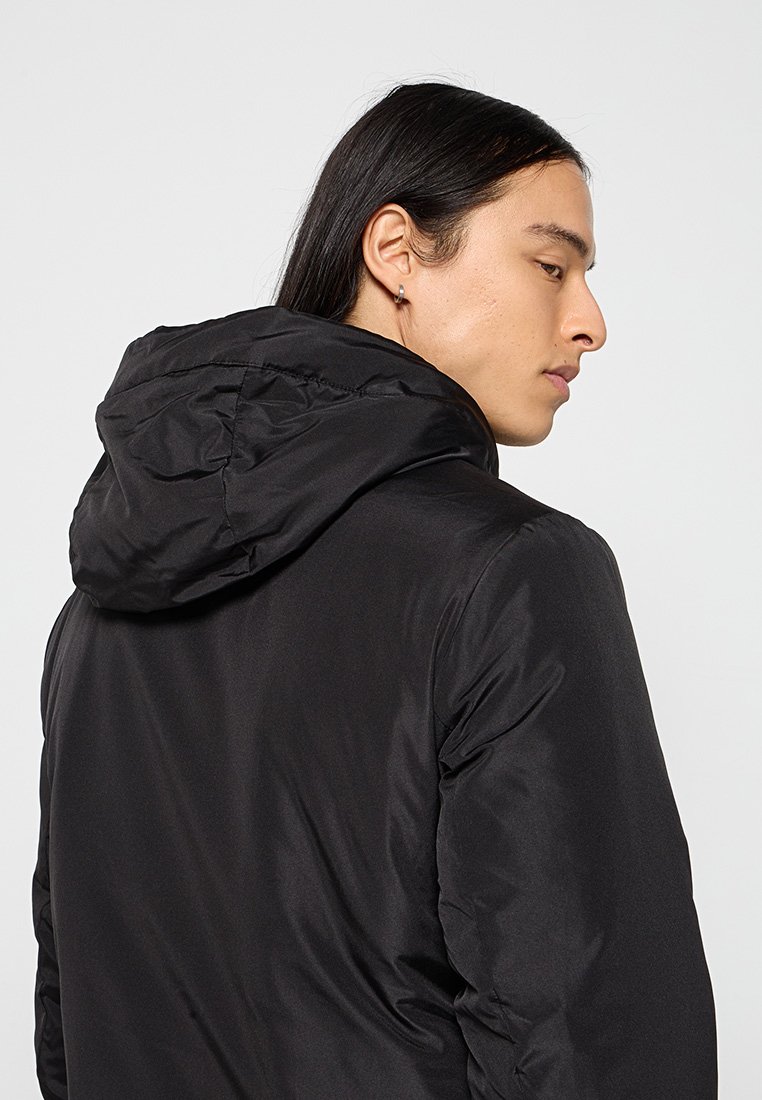 Veste doudoune noire isolée avec capuche, dotée d'une finition lisse et brillante et de détails de couture minimaux, portée par une personne vue de dos.