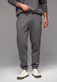 Graue Sweatpants aus weichem Stoff, mit elastischem Bund, Kordelzug, Rippbündchen und Seitentaschen. Kombiniert mit weißen Turnschuhen.