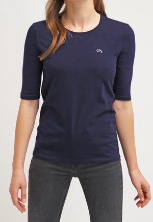 Femme portant une chemise à manches courtes bleu marine avec un petit logo blanc sur la poitrine et un jean gris foncé, debout les bras détendus.