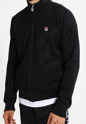 Mężczyzna w czarnej kurtce zapinanej na zamek oraz dopasowanych czarnych spodniach z naszywkami z logo, stojący na białym tle, ręce swobodnie zwisające wzdłuż ciała.