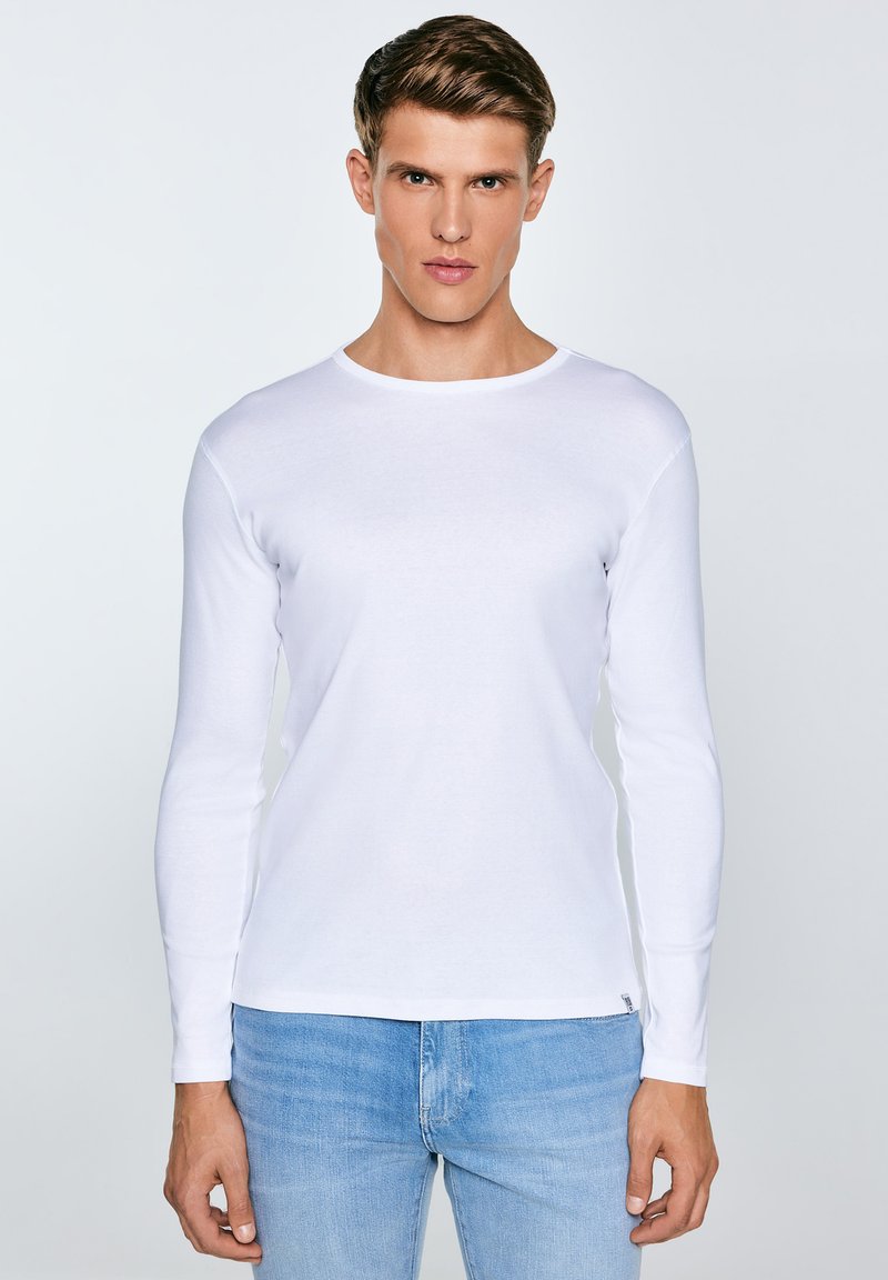 Jauns vīrietis, kas valkā vienkāršu baltu garām piedurknēm kreklu un gaiši zilas džinsas, stāvot pret vienkāršu gaiši pelēku fonu.