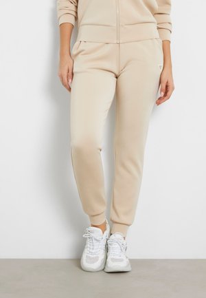 Beige jogger pants made of smooth fabric with a tapered design. Features elasticated cuffs and subtle side pockets. Worn with white sneakers.