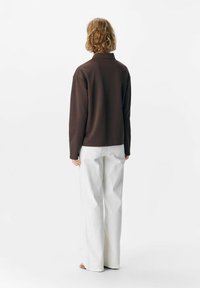 Braunfarbener Langarmoberteil mit hohem Kragen, kombiniert mit weißen weiten Hosen. Der Stoff wirkt weich und leicht strukturiert. Minimalistisches Design.
