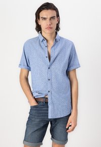 Chemise à manches courtes bleu clair avec un motif subtil, assortie à un short en denim foncé. La chemise possède un col pointu et des manches retroussées.