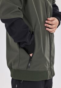 Oliven grøn zip-up jakke med en sort ærme, ribbet manchet og en sidelomme med lynlås. Glat stoftekstur med subtile syninger og logo.