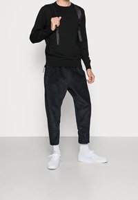 Černý svetr s dlouhým rukávem, texturované tmavě šedé manšestrové kalhoty se zúženými nohavicemi, doplněné bílými teniskami a bílými kotníkovými ponožkami.