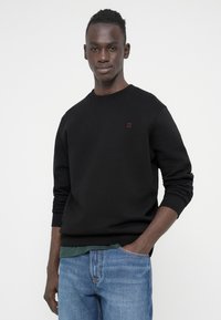 Juodas džemperis su apvalia iškirpte, ribbintomis rankogaliais ir mažu raudonu "II" logotipu ant krūtinės, derinamas su mėlynais džinsais ir žaliu apatinio marškinėlio sluoksniu.