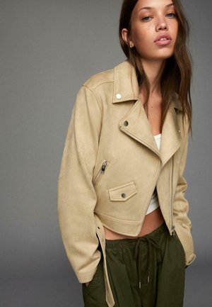 Korte beige suède motorjack met een cropped ontwerp, ritszakken en zilverkleurige hardware-accenten, gecombineerd met groene broek met trekkoord.