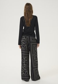Schwarzes langärmeliges Oberteil kombiniert mit weit geschnittenen Hosen, die ein geometrisches Muster in Gold zeigen, und hebt einen lockeren Schnitt sowie einen strukturierten Stoff hervor.