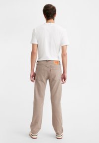 Los jeans de mezclilla beige tienen un corte recto y bolsillos traseros. La cintura incluye una etiqueta de marca amarilla. El modelo lleva una camiseta blanca lisa.