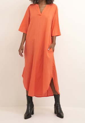 Langes oranges Kleid aus leichtem Stoff, mit V-Ausschnitt, 3/4-Ärmeln und seitlichen Schlitzen. Getragen mit schwarzen Knöchelstiefeln.