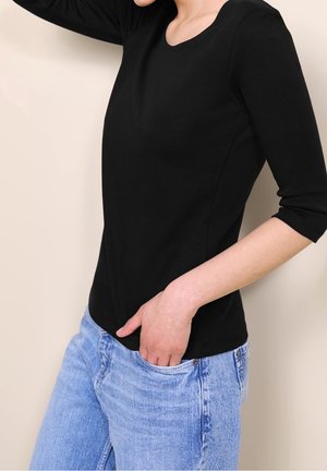 Persona con una camiseta ajustada negra de manga tres cuartos y jeans azul claro, con una mano en el bolsillo delantero.