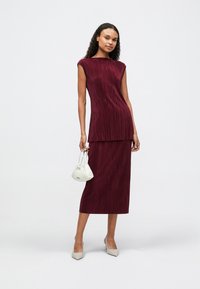 Σέτ με πλισέ τοπ σε μπορντό απόχρωση και midi φούστα με ίσια σιλουέτα, συμπληρωμένο με άσπρη τσάντα χεριού και μπεζ γόβες.