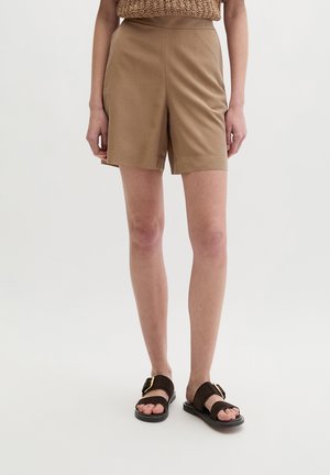 Osoba ubrana w beżowe szorty z wysokim stanem i ciemnobrązowe sandały ze złotymi klamrami, stojąca na tle jednolitego jasnego tła.