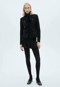 O ținută din materiale mixte de culoare neagră, care include o jachetă ajustată, cu nasturi și buzunare, și o fustă cu tiv ondulat, asortată cu colanți negri și pantofi cu toc lustruiți.